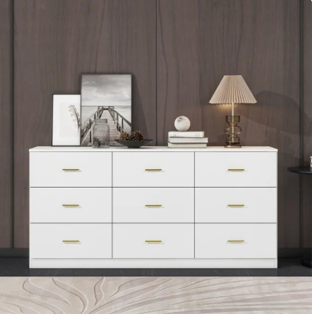 Modern white 9-drawer dresser with gold handles, placed in a contemporary room setting with decorative items on top against a dark wood wall.